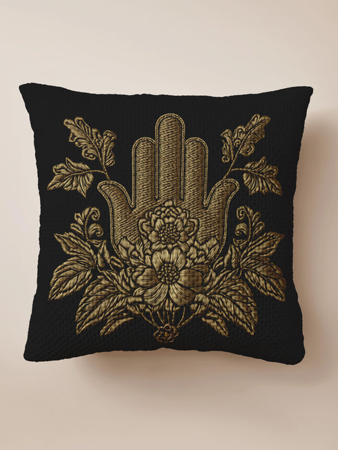 A black woven throw pillow featuring an embroidered gold Hamsa hand surrounded by intricate floral and botanical designs.